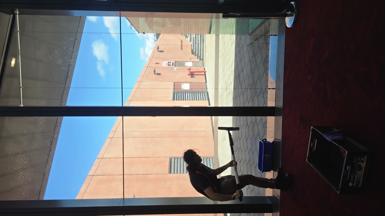 A woman cleaning the floor in a building lobby with a squeegee. Outside, a brick building and blue sky are visible through large glass windows.