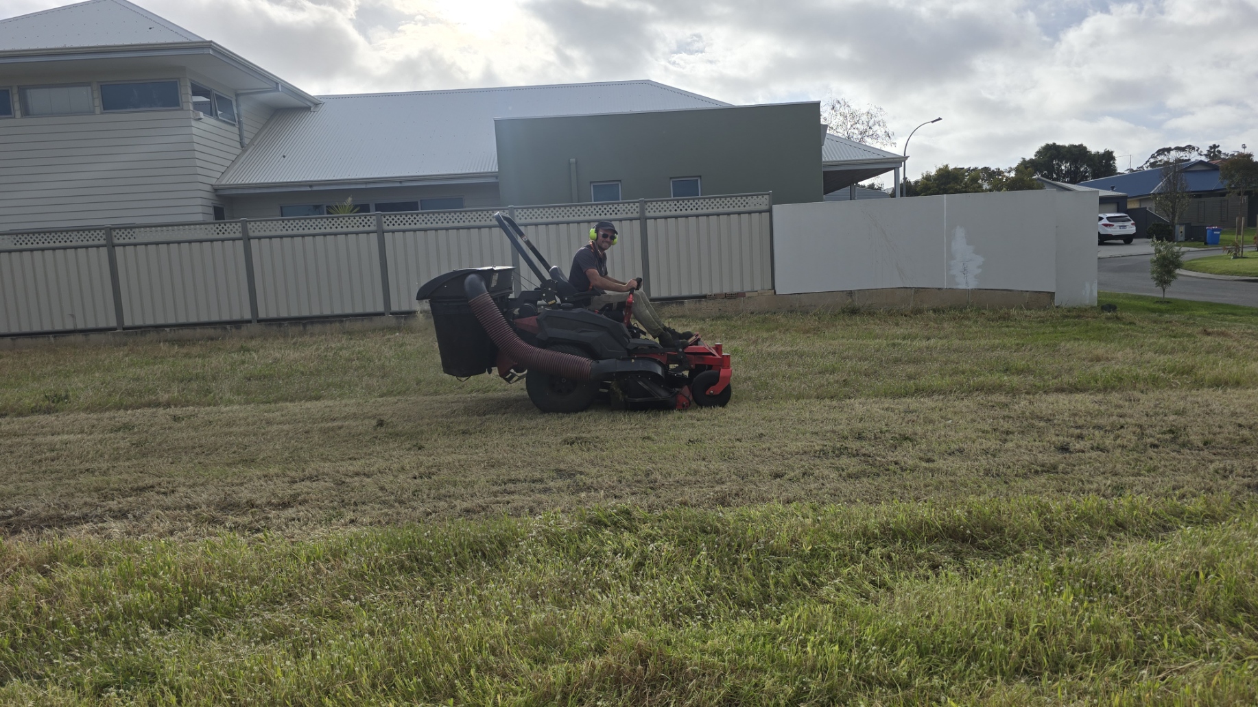 A man with headphones riding a riding lawn mower on a grassy field in a residential neighborhood with houses, a fence, trees, and a cloudy sky in the background.