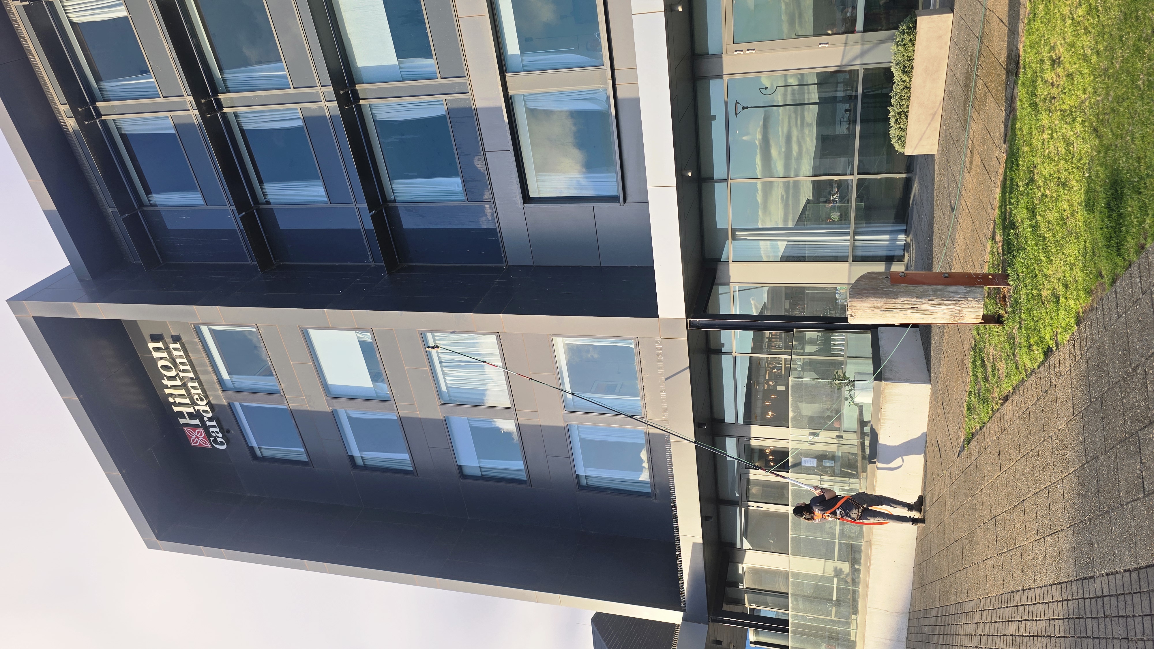 The image shows a modern multi-story hotel building with a sign that reads 'Hilton Garden Inn' near the top. In front of the hotel, a person is using a cleaning tool connected to a long pole, reaching up to the top of the building. The building has large windows with curtains, and the bottom floor appears to have a lobby area with glass doors and windows. The surrounding area includes a paved sidewalk, some green grass, and a few street lamps.