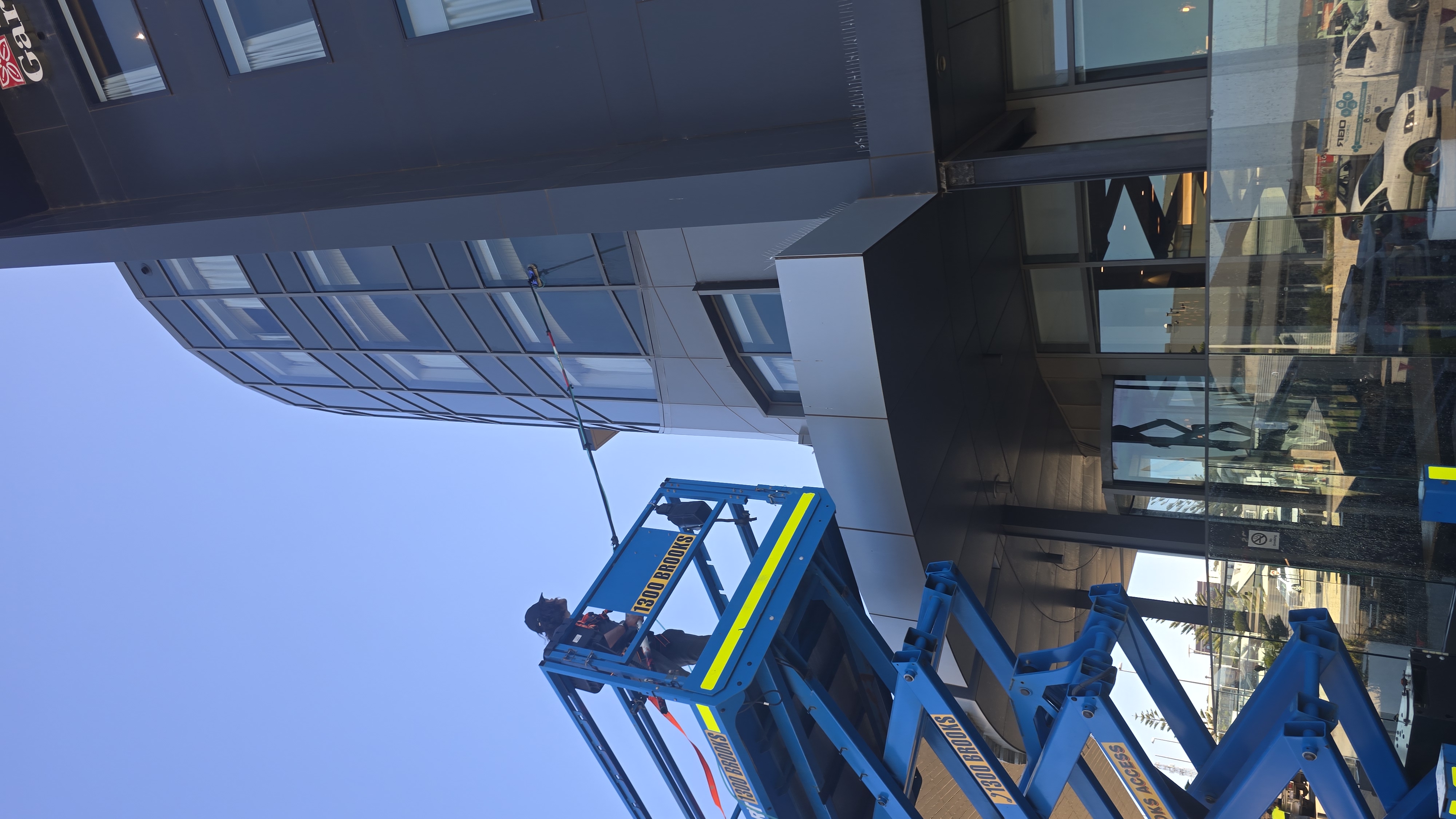 A worker on a blue hydraulic lift cleaning the exterior windows of a tall modern building with a curved glass facade and dark exterior panels.