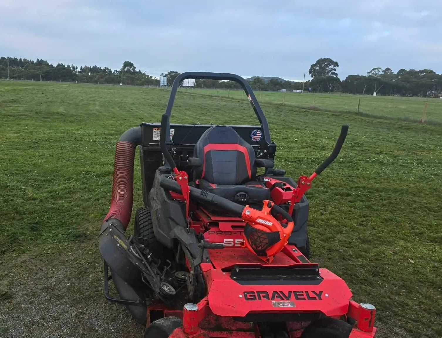 Red and black Gravely lawn mower with seat, grass collection system, and a blower, on a grassy field with trees and cloudy sky in the background.