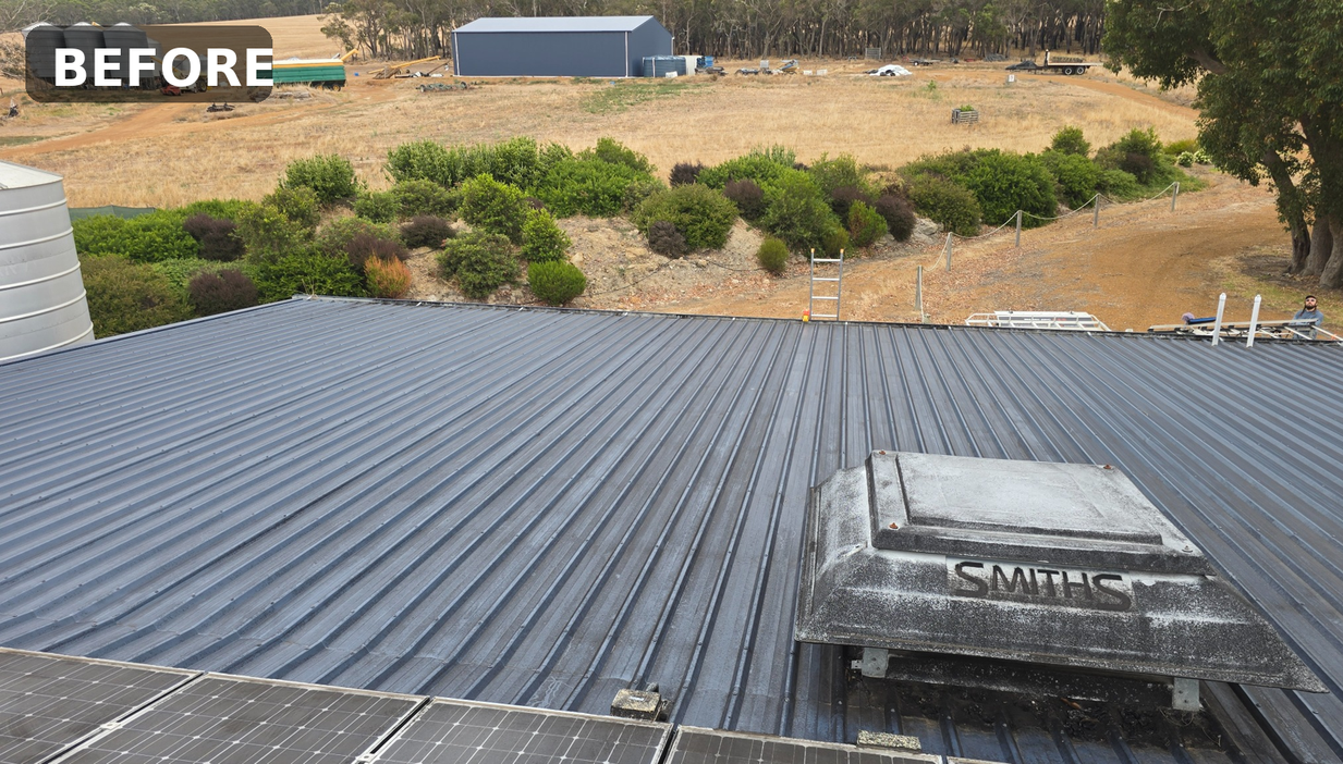 View of a metal roof with a ventilation unit labeled "SMITHS" on top, and solar panels in the bottom left corner. In the distance, there are trees, bushes, a dirt path, and farm buildings.